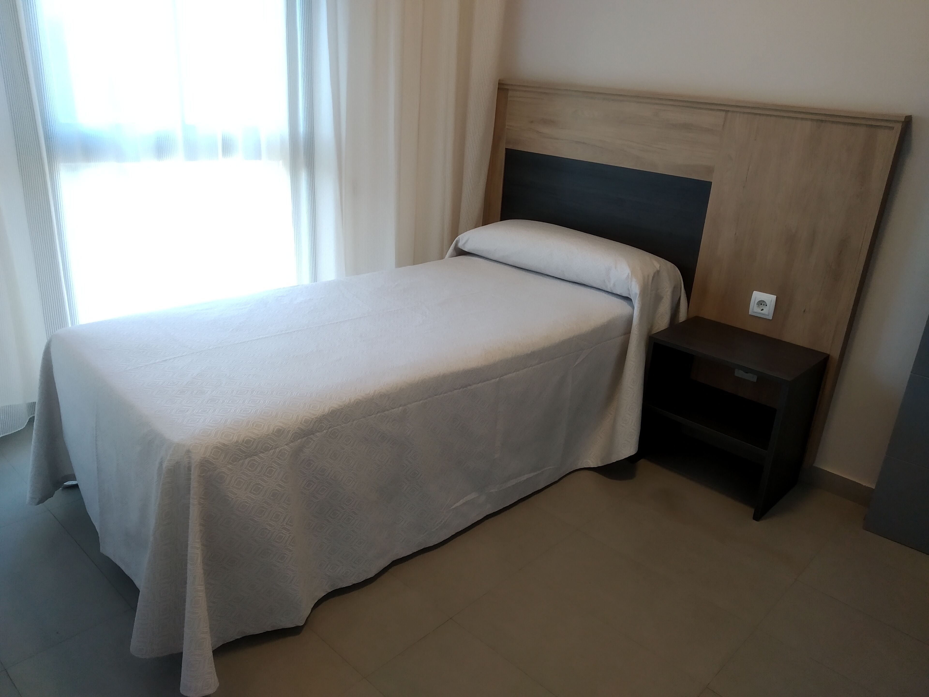 A single bed with a wooden headboard, a bedside table, and a power outlet on the wall.