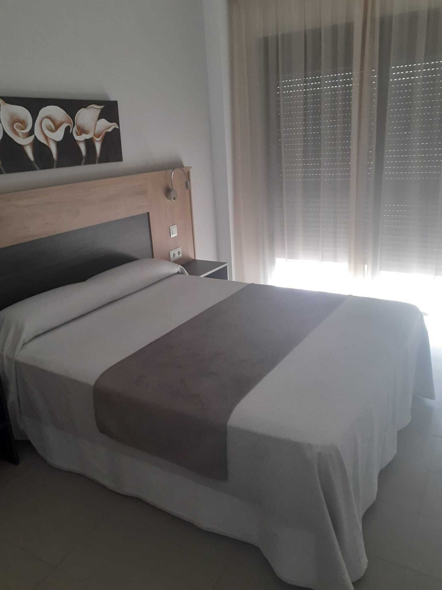 A neatly made bed with a wooden headboard, a bedside table, and a window with sheer curtains.