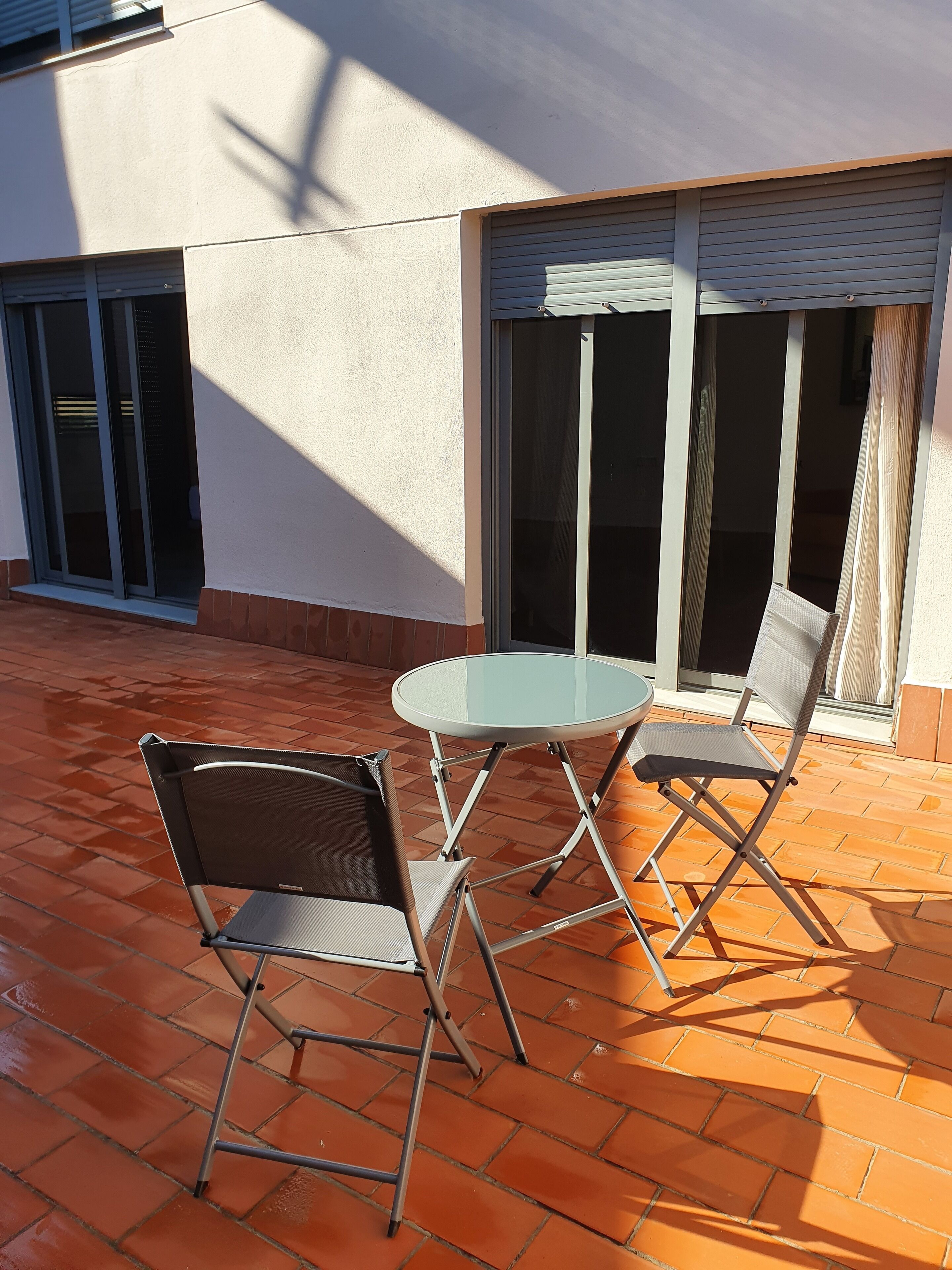 An outdoor patio with a round glass table and two foldable chairs, a sliding glass door, and a tiled floor.