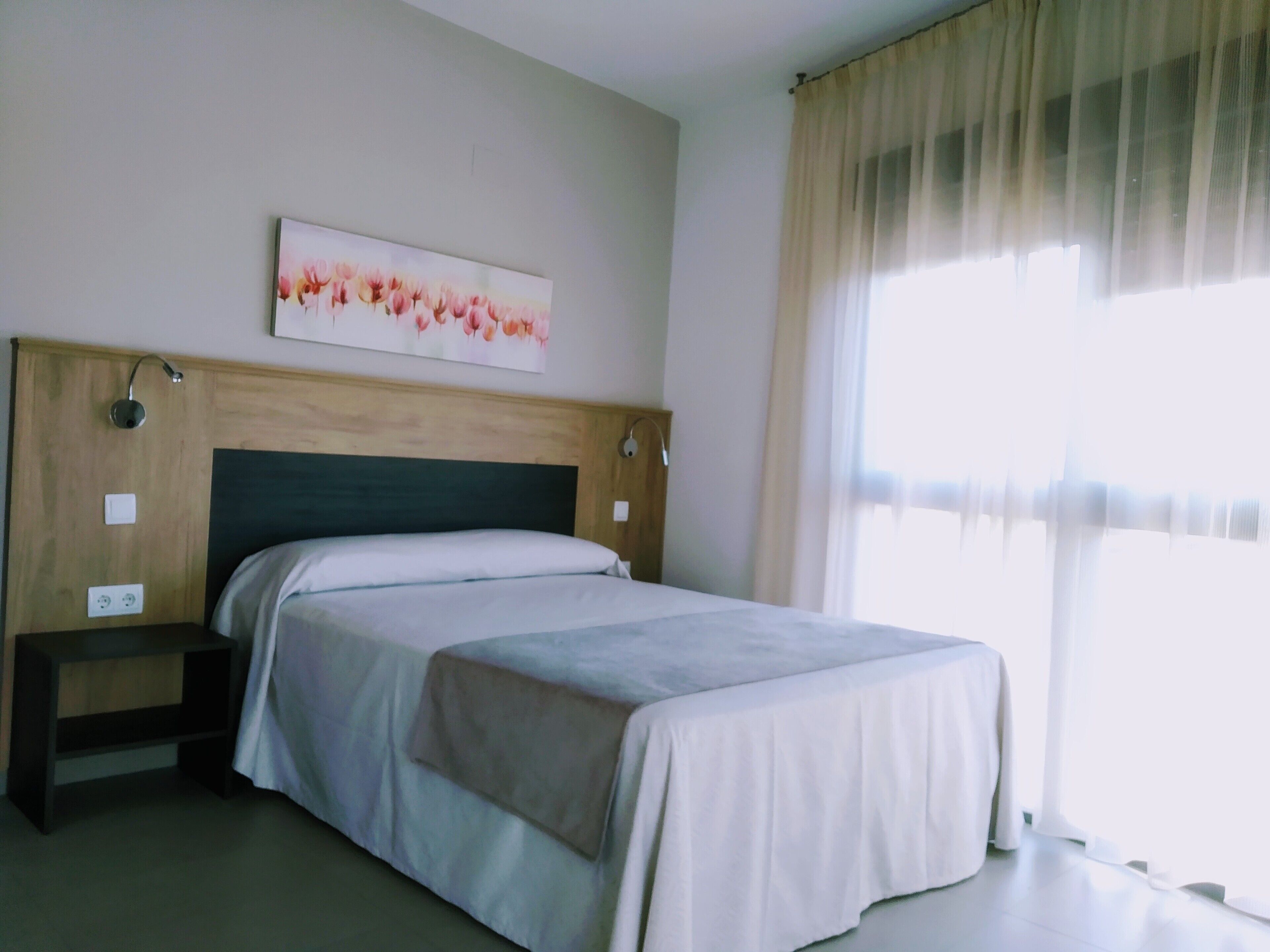 A hotel room with a bed, a nightstand, a wall-mounted lamp, a painting, and a window with curtains.