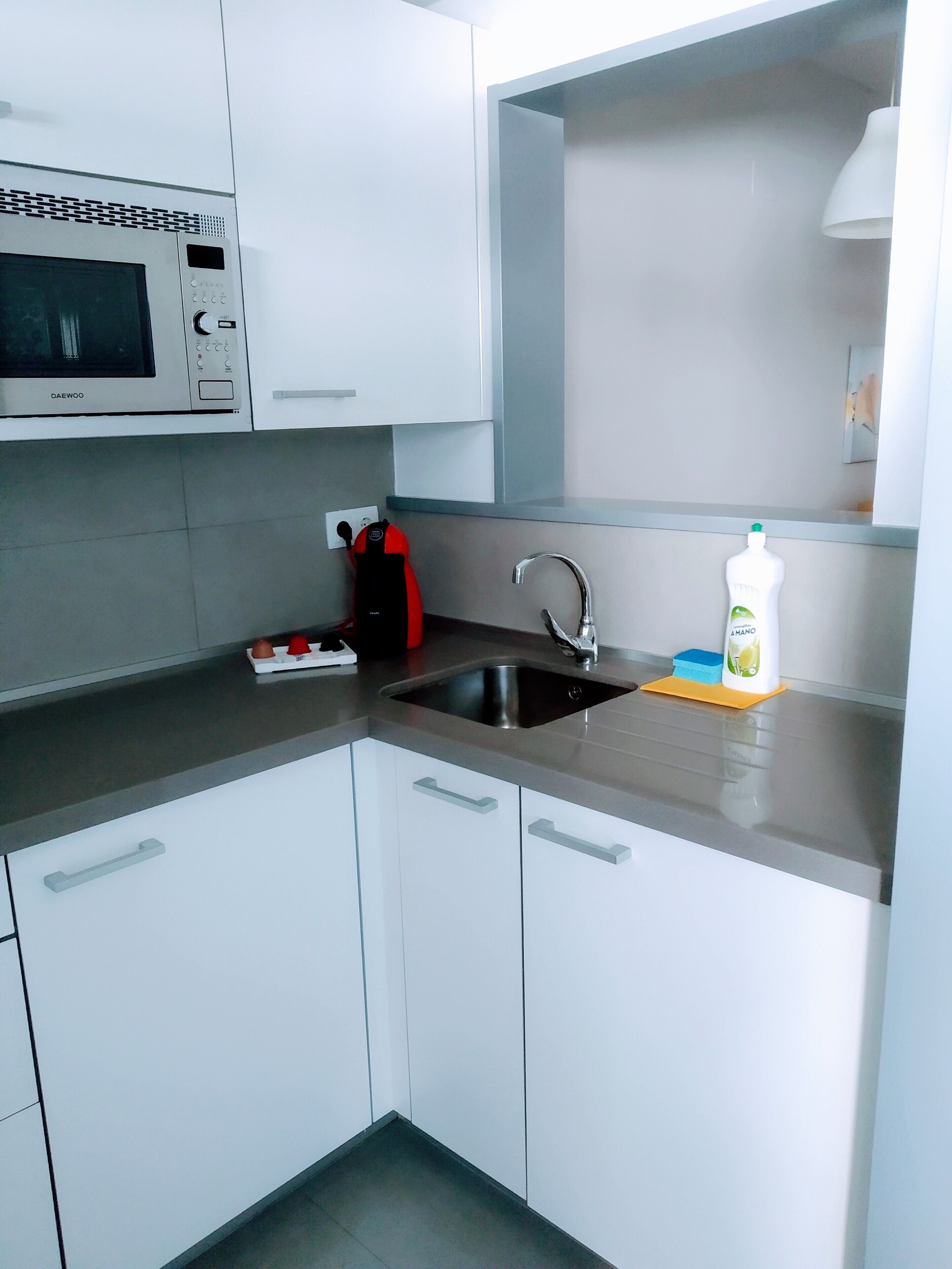 Une cuisine moderne avec des meubles blancs, un évier en acier inoxydable, un four à micro-ondes et un flacon de liquide vaisselle.