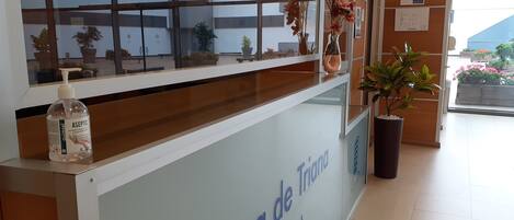 Un mostrador de recepción con vista despejada al exterior, un dispensador de gel hidroalcohólico y una planta en maceta.