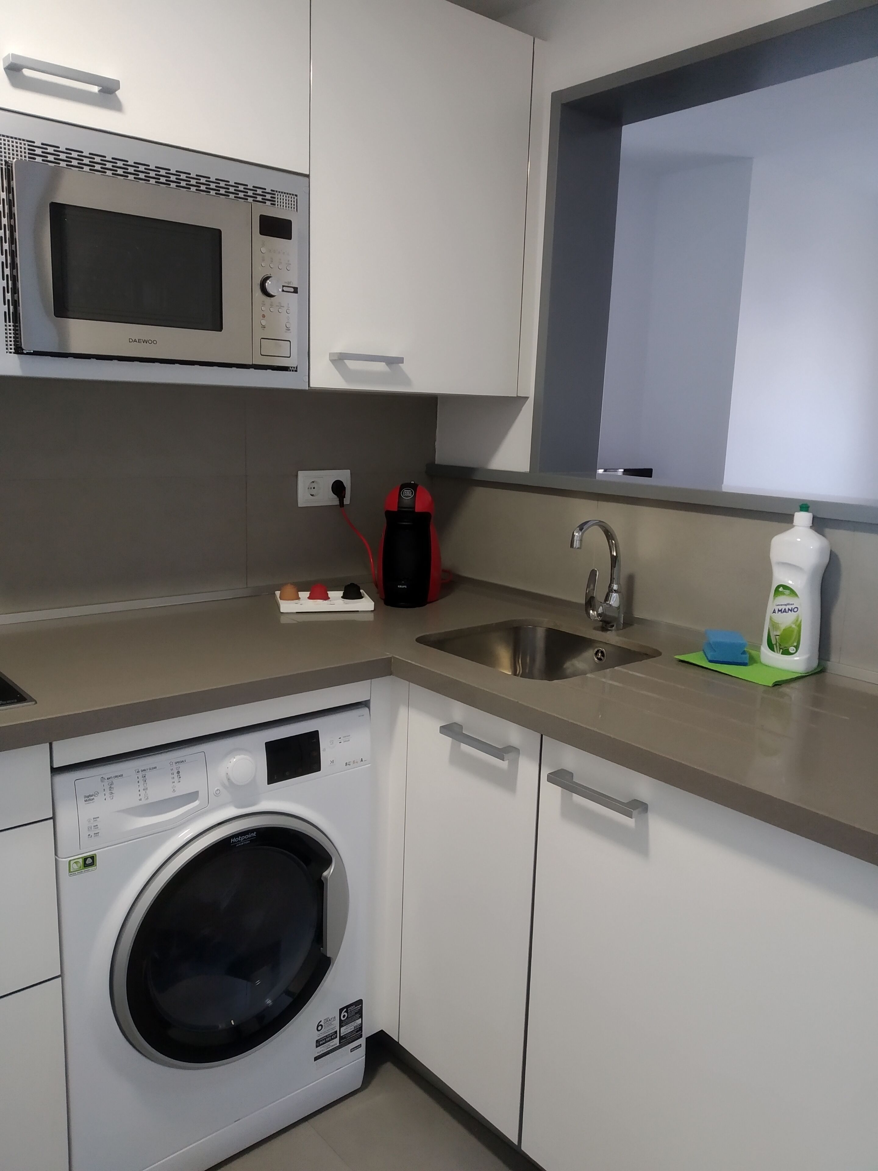A compact kitchen with a microwave, a washing machine, a sink, and a window above the sink.