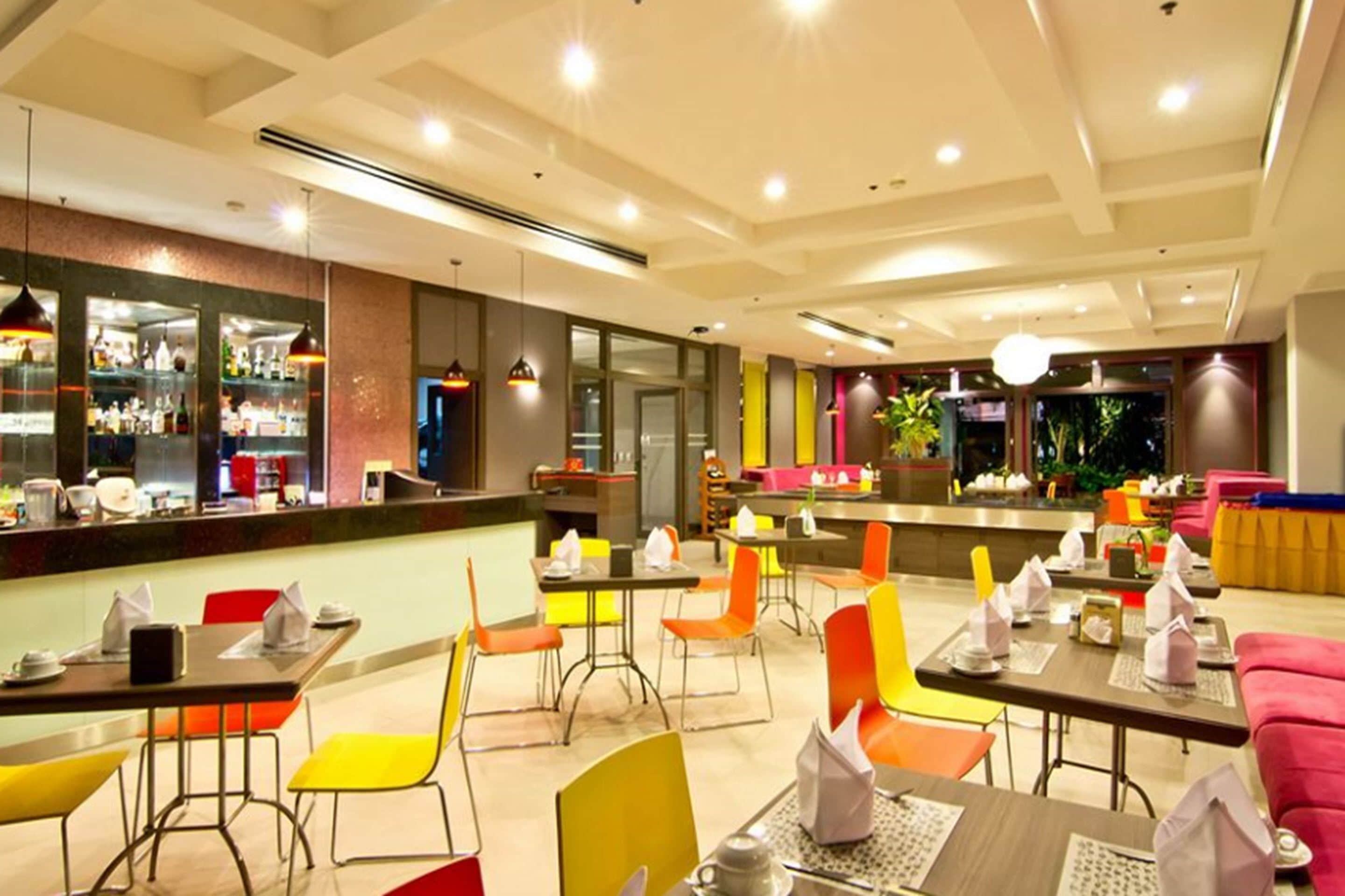 A modern restaurant with bright orange and yellow chairs, wooden tables, and a bar area with shelves of bottles.