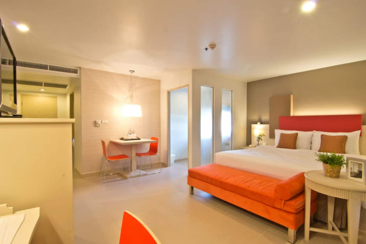 Ein modernes Hotelzimmer mit einem großen Bett, einem Schreibtisch mit zwei Stühlen, einem kleinen Tisch mit Pflanze und einem Spiegel.