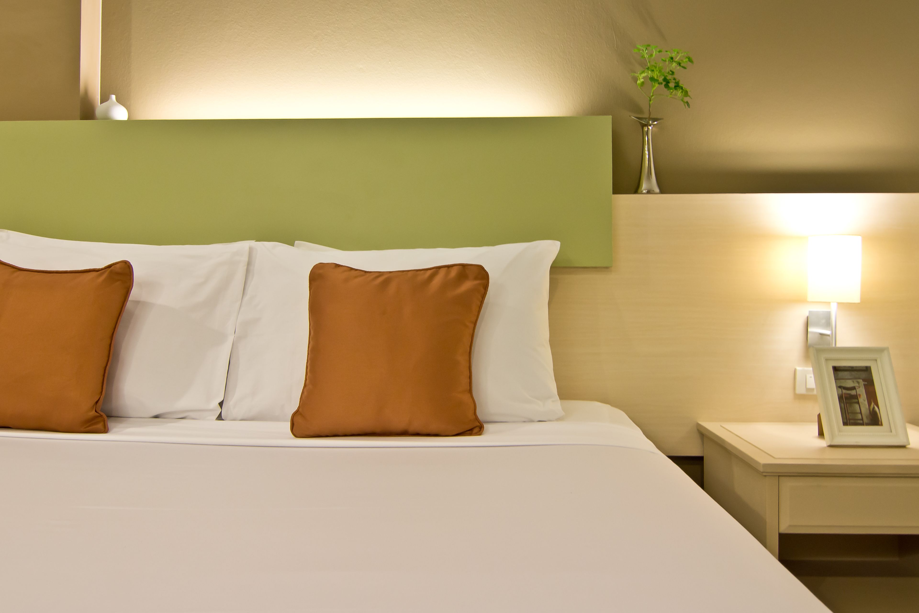 A neatly made bed with white and brown pillows, a bedside table with a framed picture, and a wall-mounted vase with a plant.