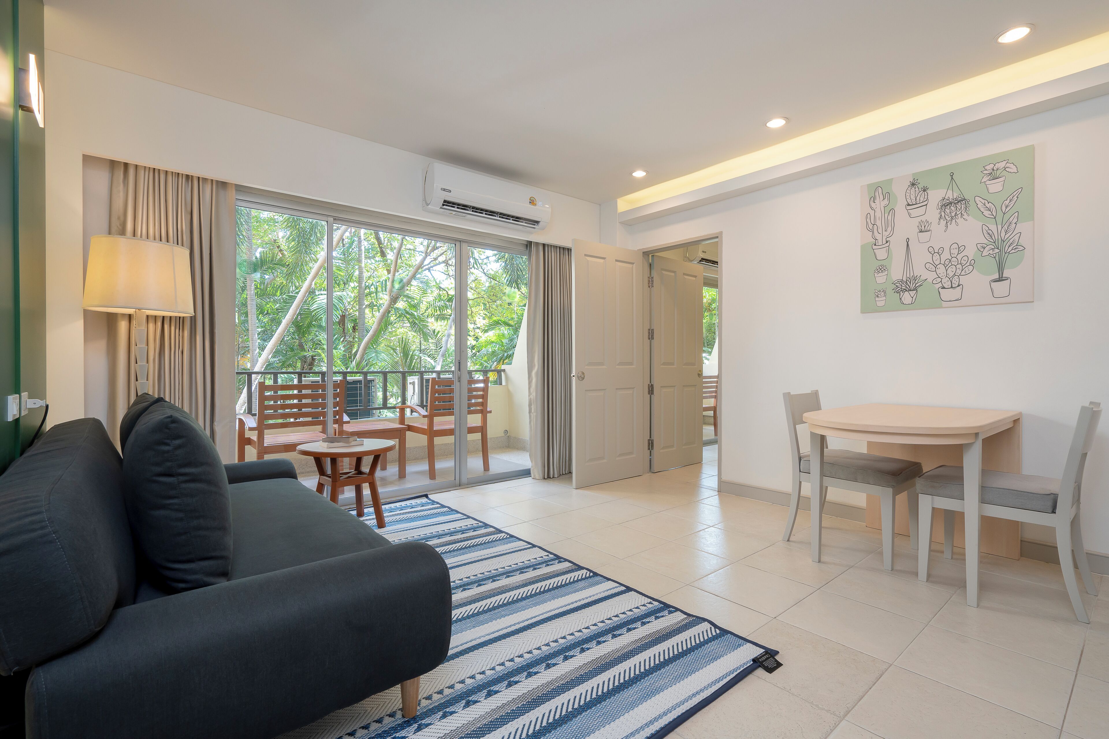 A modern living room with a sofa, a dining table, and chairs. There is a large window with curtains, an air conditioning unit, and a wall-mounted artwork.