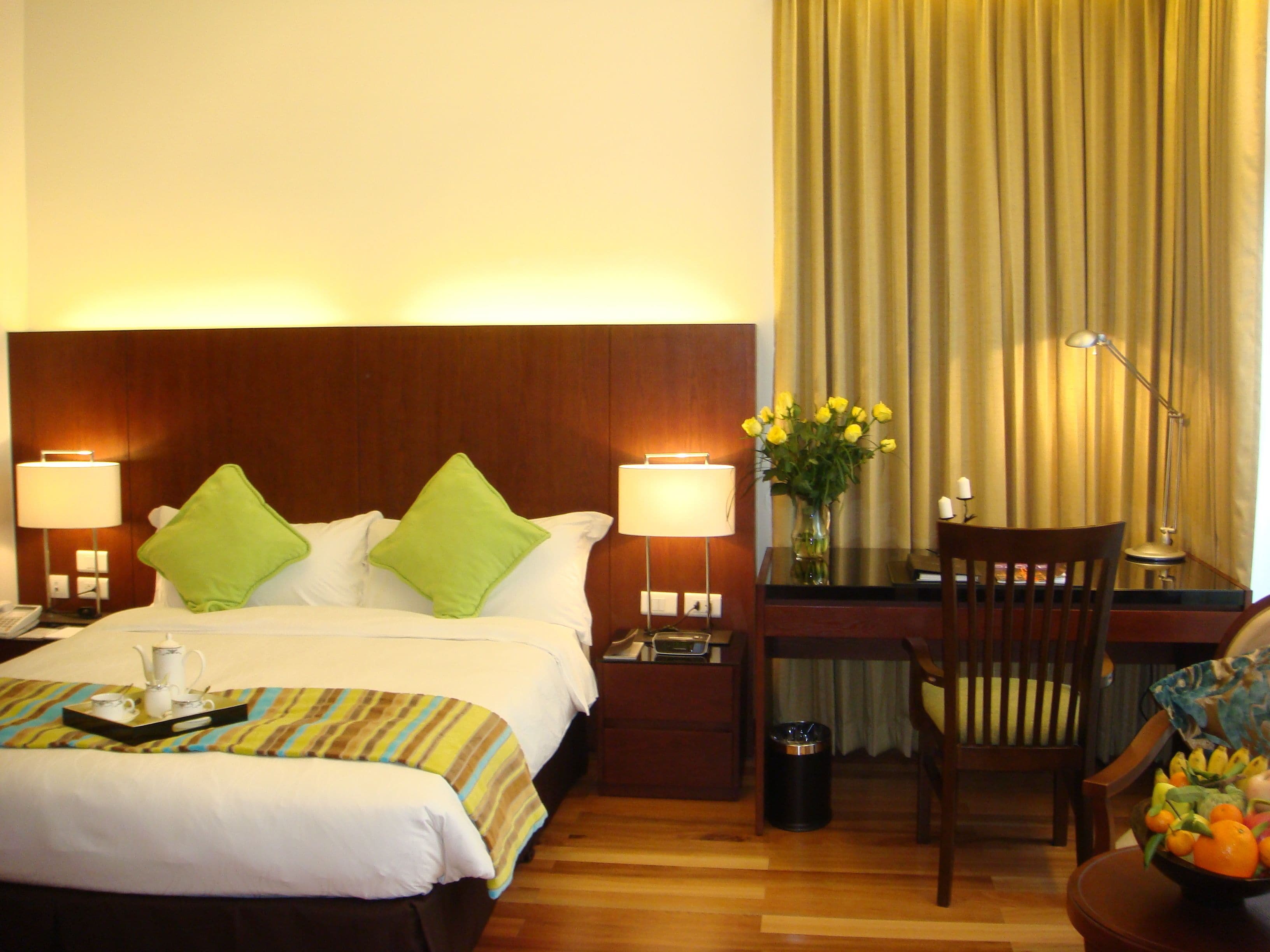Une chambre d’hôtel comprenant un lit, des lampes de chevet, un bureau avec une chaise, un vase avec des fleurs et un panier de fruits.