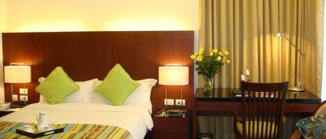 A hotel room with a bed, bedside lamps, a desk with a chair, a vase with flowers, and a fruit bowl.