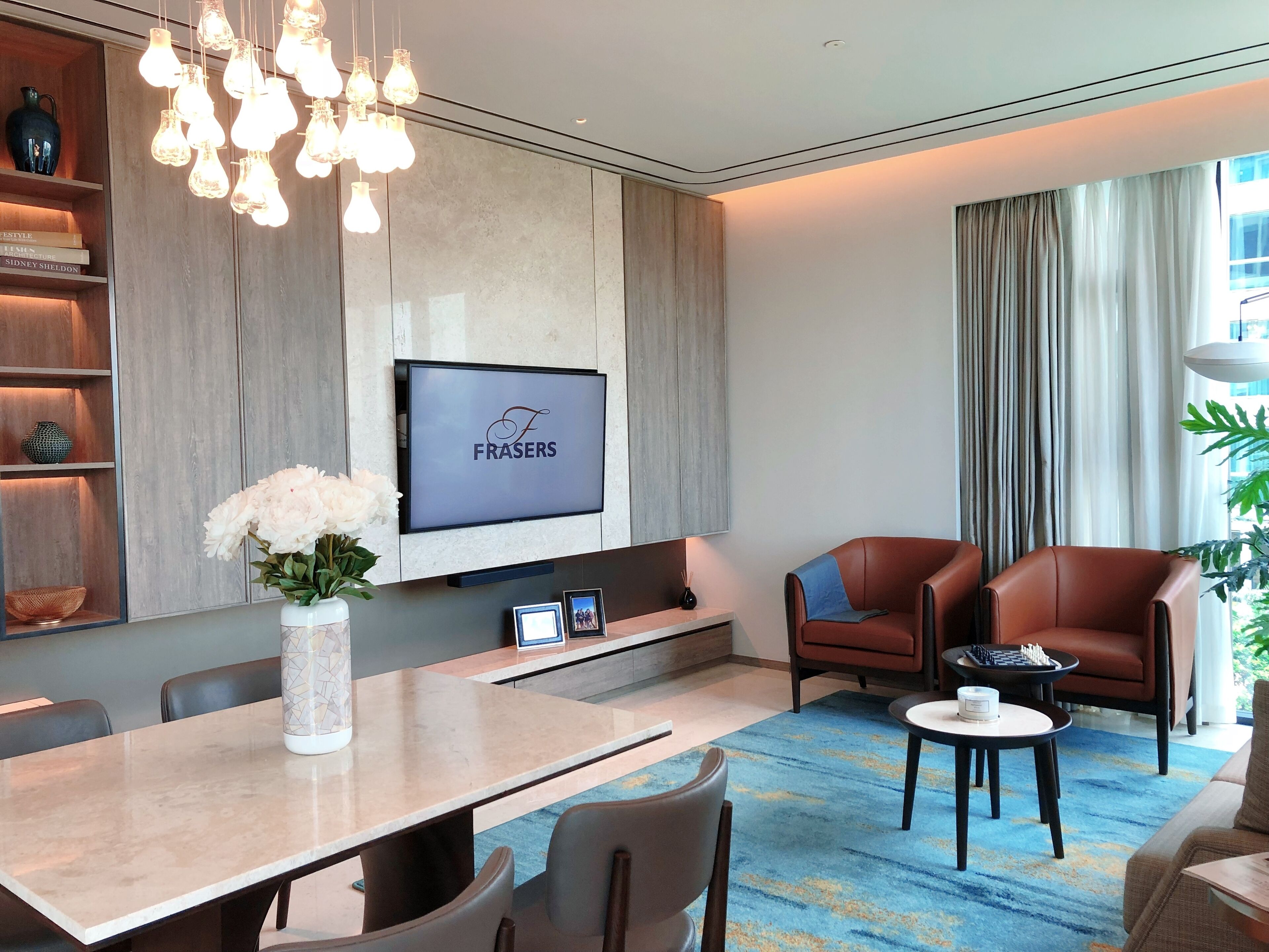 Una habitación de hotel moderna con televisión de pantalla plana, un comedor con mesa y sillas, un área de estar con dos sillones de cuero y un ventanal con cortinas.