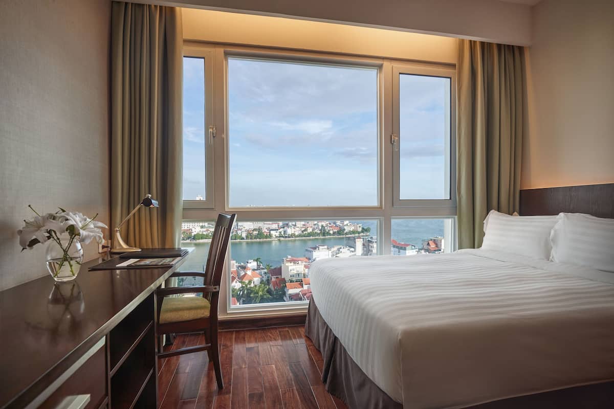 A hotel room with a large bed, a desk with a chair, a vase of flowers, and a view of a city and water through large windows.