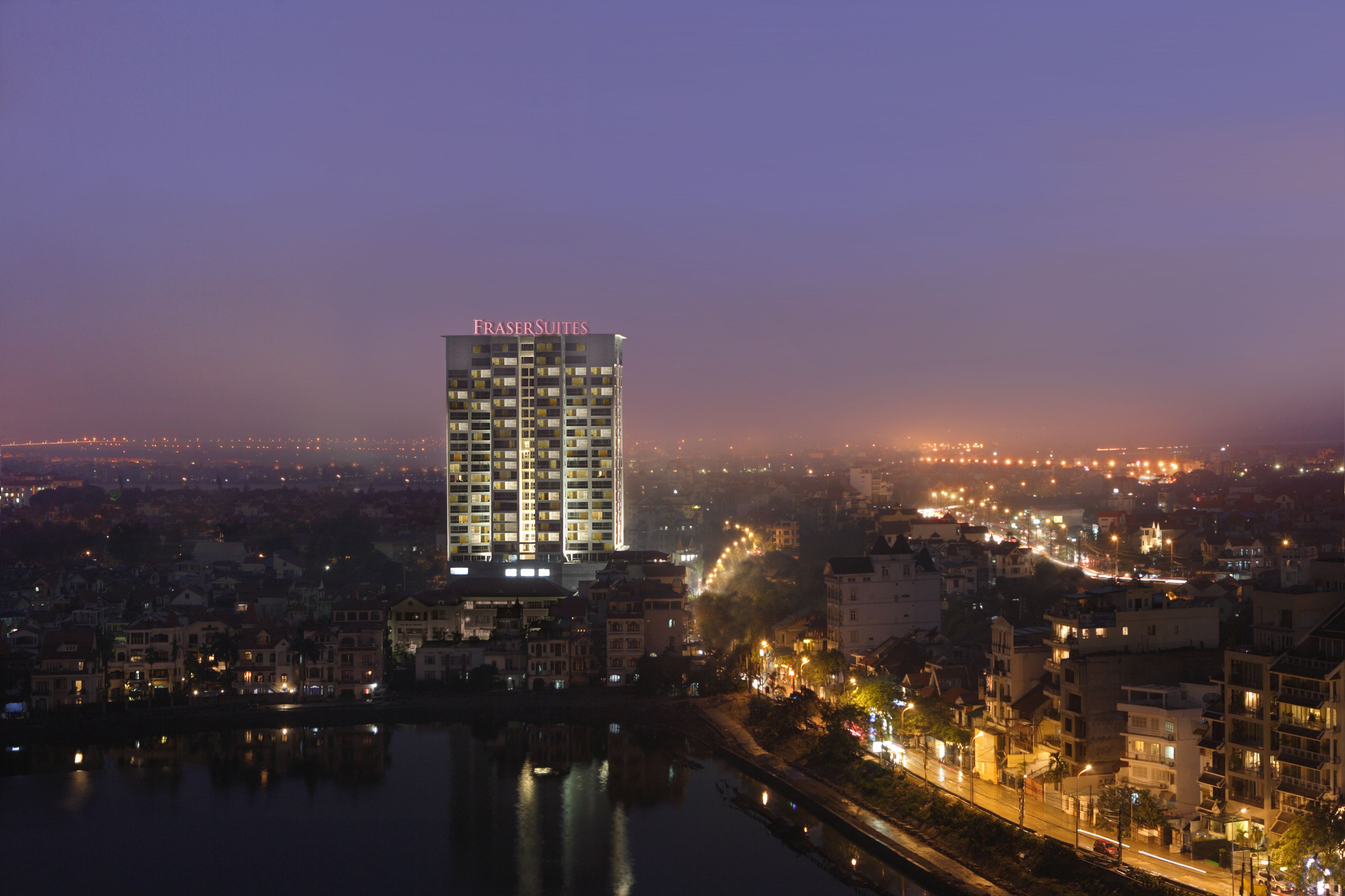 Un hotel moderno de gran altura con un letrero llamativo, ubicado junto a la costa con luces de la ciudad al fondo.