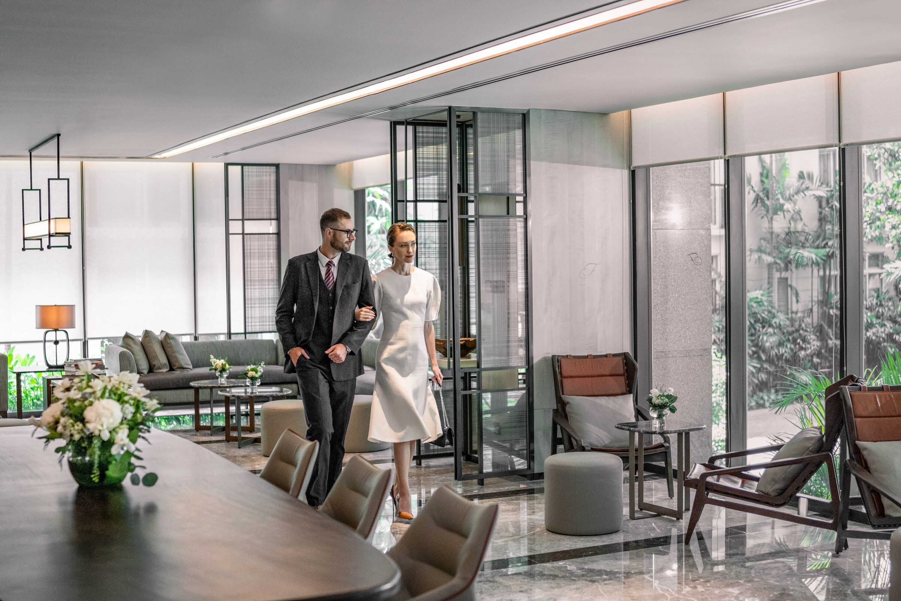 A modern hotel lobby with a large dining table, comfortable seating, and large windows offering a view of greenery.