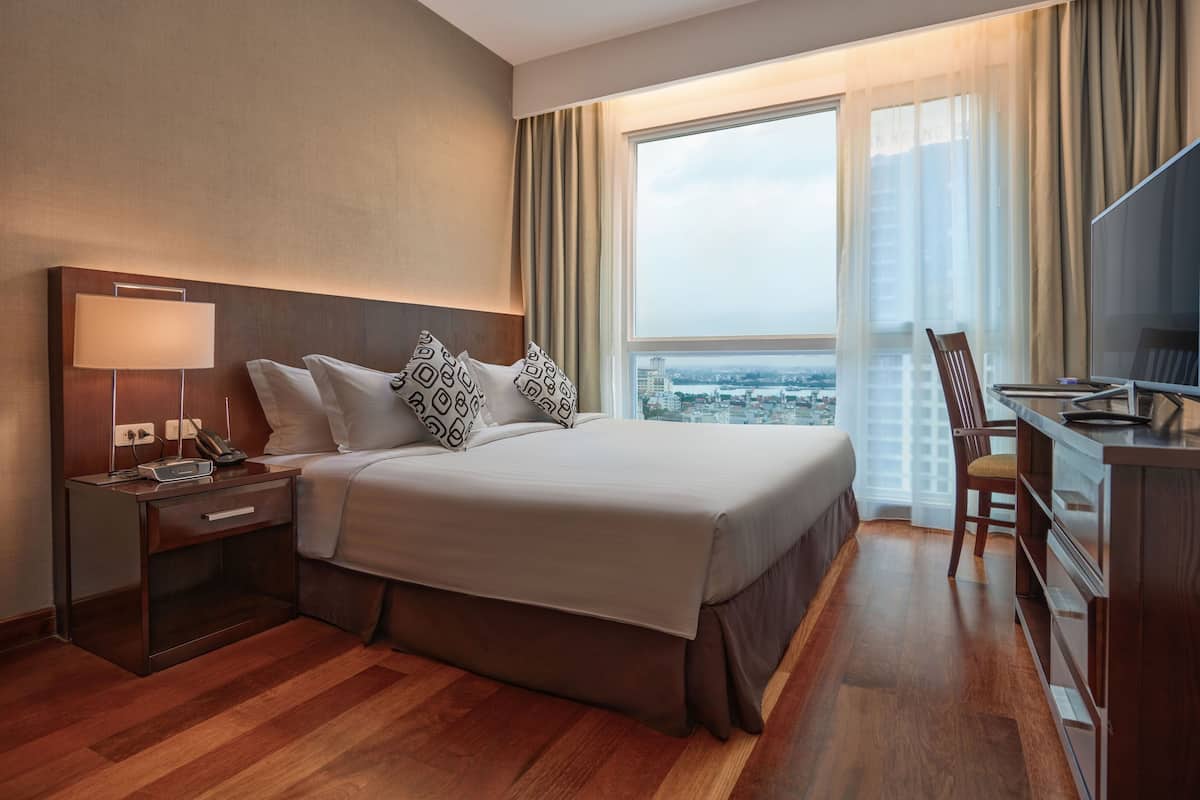 A hotel room with a large bed, a desk with a TV, a nightstand, and a view of the city through large windows.