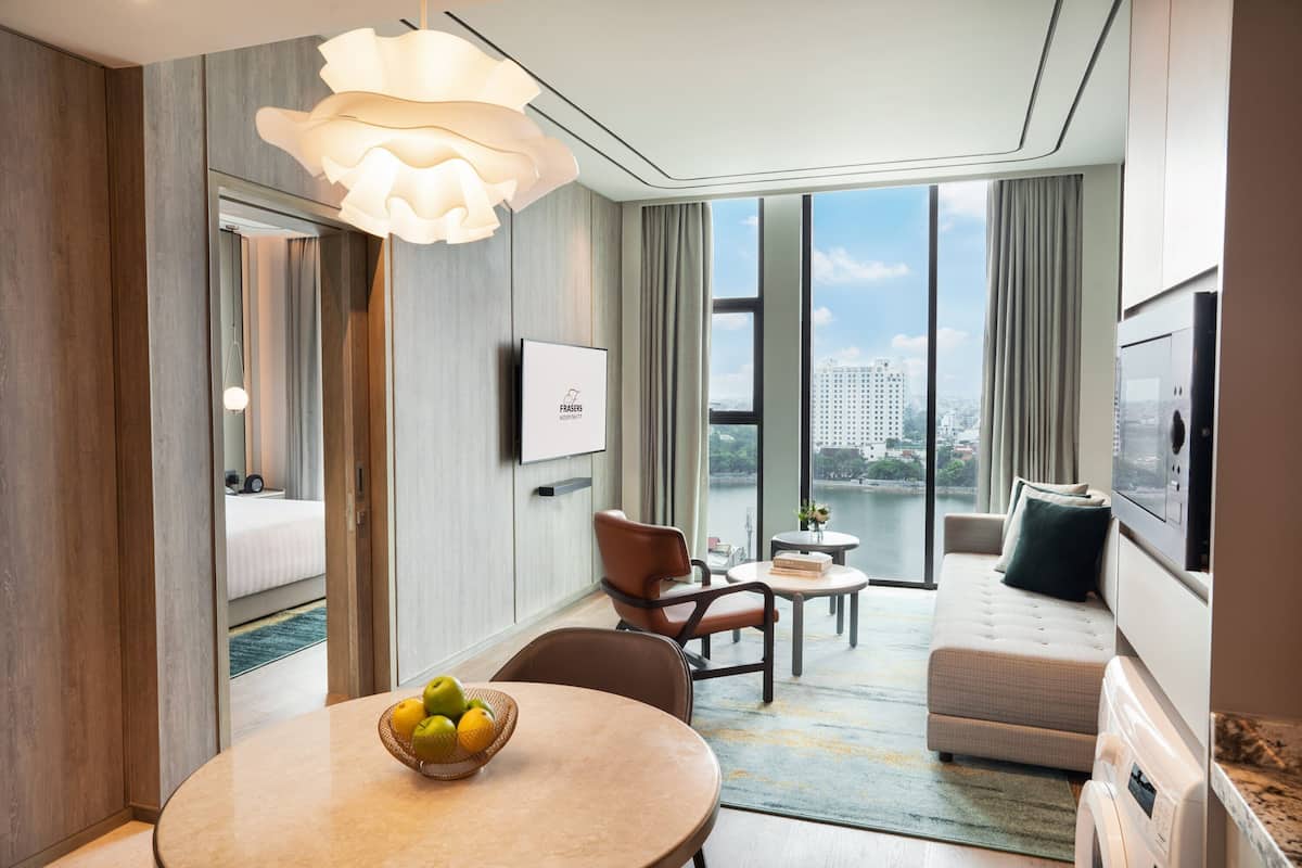 A modern hotel room with a large window, a round dining table with fruit, a sofa, and a TV.