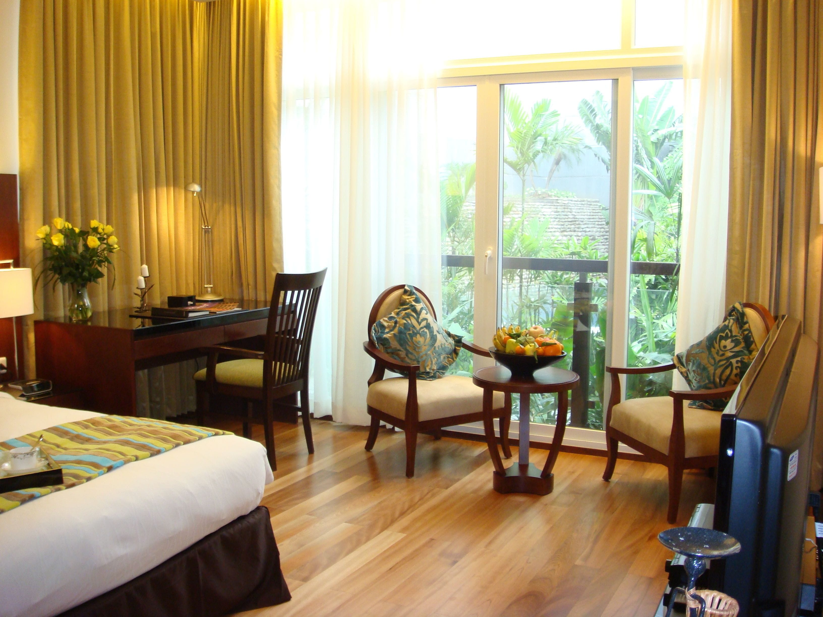 A hotel room with a bed, desk, two chairs, a small table with fruit, and a view of greenery through large windows.