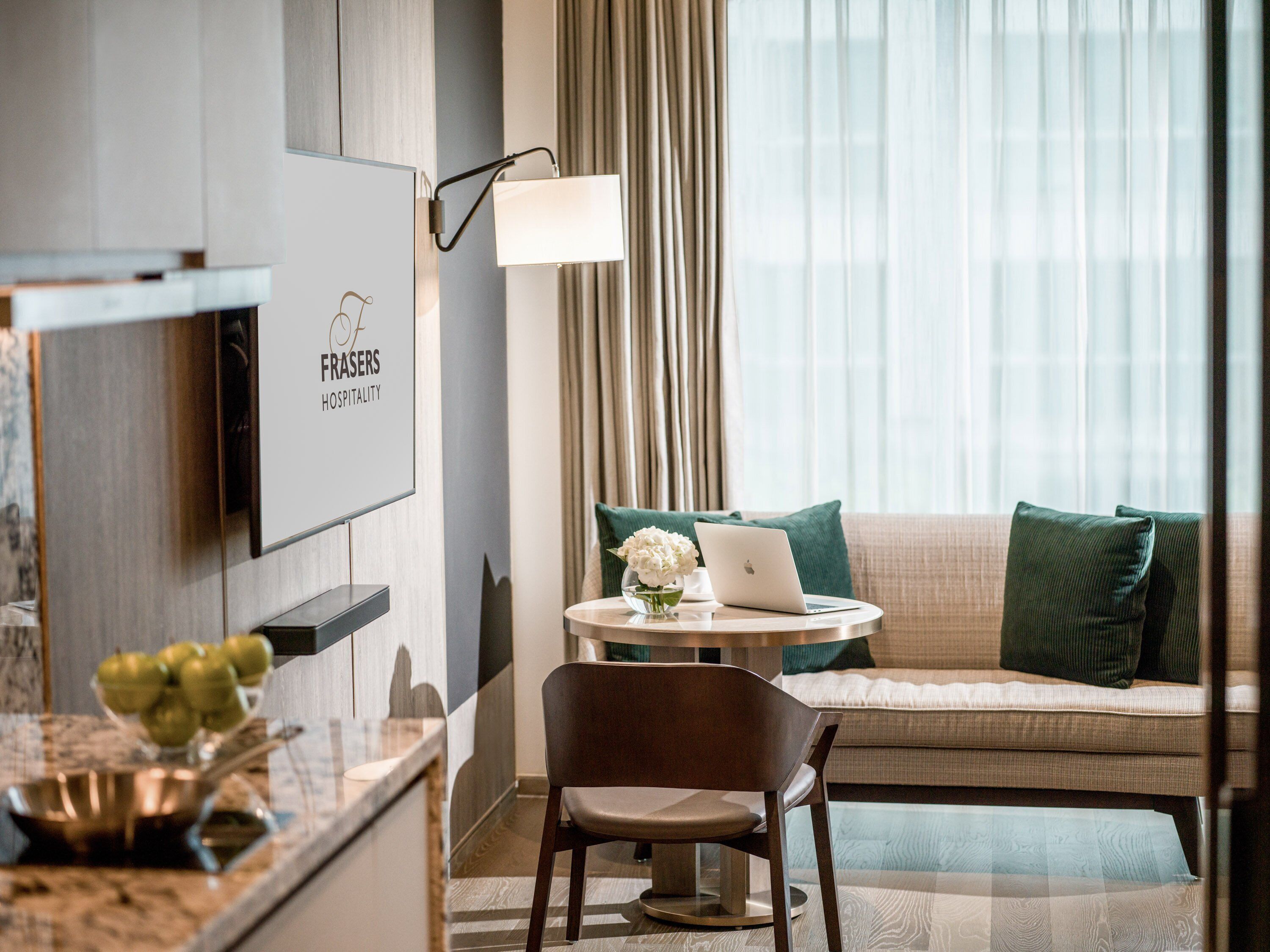 A modern hotel room with a sofa, a dining table with a laptop, a TV mounted on the wall, and a decorative bowl with apples.
