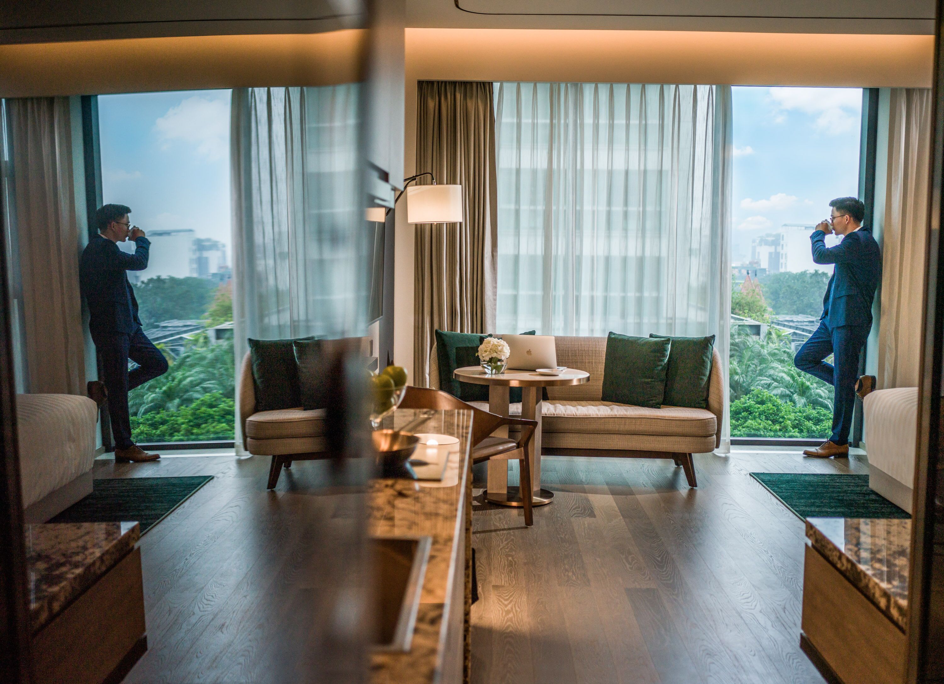 Une chambre d’hôtel moderne avec un canapé, une table basse et un ordinateur portable posé sur une petite table. De grandes fenêtres, équipées de rideaux, offrent une vue sur la verdure et les bâtiments environnants.