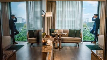 Une chambre d’hôtel moderne avec un canapé, une table basse et un ordinateur portable posé sur une petite table. De grandes fenêtres, équipées de rideaux, offrent une vue sur la verdure et les bâtiments environnants.
