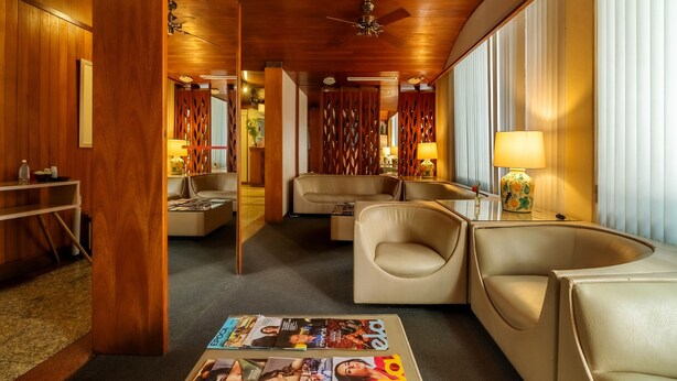 A hotel room with a wooden ceiling, a ceiling fan, a sofa, a chair, a table with magazines, and a lamp.