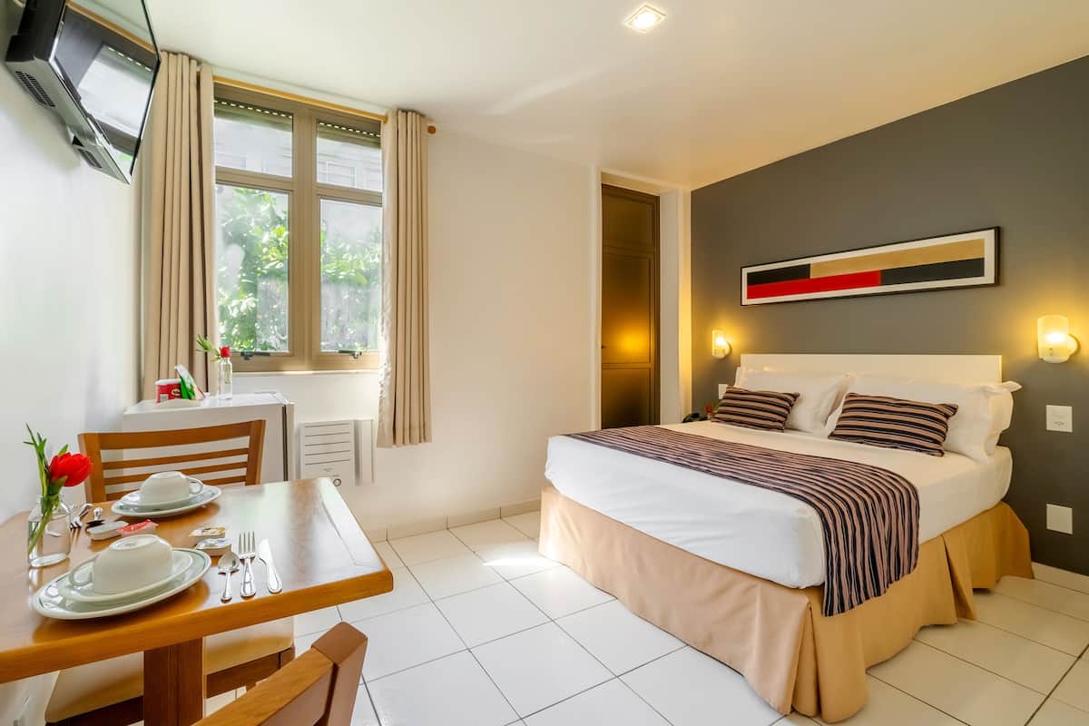 Une chambre d’hôtel avec un lit, une table à manger avec des assiettes et des couverts, un téléviseur fixé au mur et une fenêtre avec des rideaux.