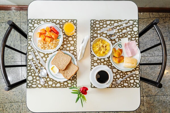 Uma mesa de café da manhã com dois pratos, cada um contendo torradas, ovos mexidos e uma fatia de presunto. Há também tigelas com frutas e um copo de suco de laranja.