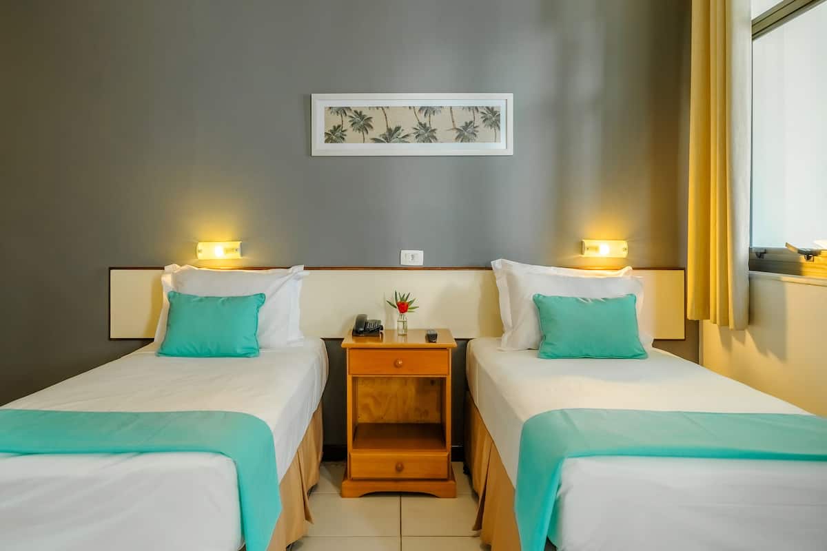 Une chambre d’hôtel avec deux lits, chacun recouvert d’une couvre-lit turquoise et agrémenté d’oreillers blancs. Une table de chevet en bois se trouve entre les deux lits, avec un téléphone et un petit vase dessus. Un tableau encadré est accroché au mur, au-dessus des lits.