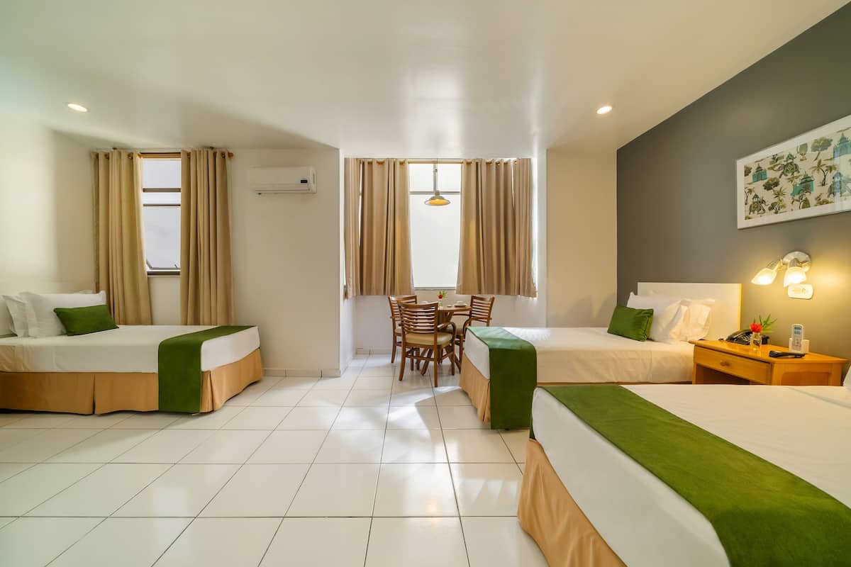 Une chambre d’hôtel avec deux lits, une table à manger, des chaises, une petite table avec un téléphone et une lampe, et une fenêtre avec des rideaux.