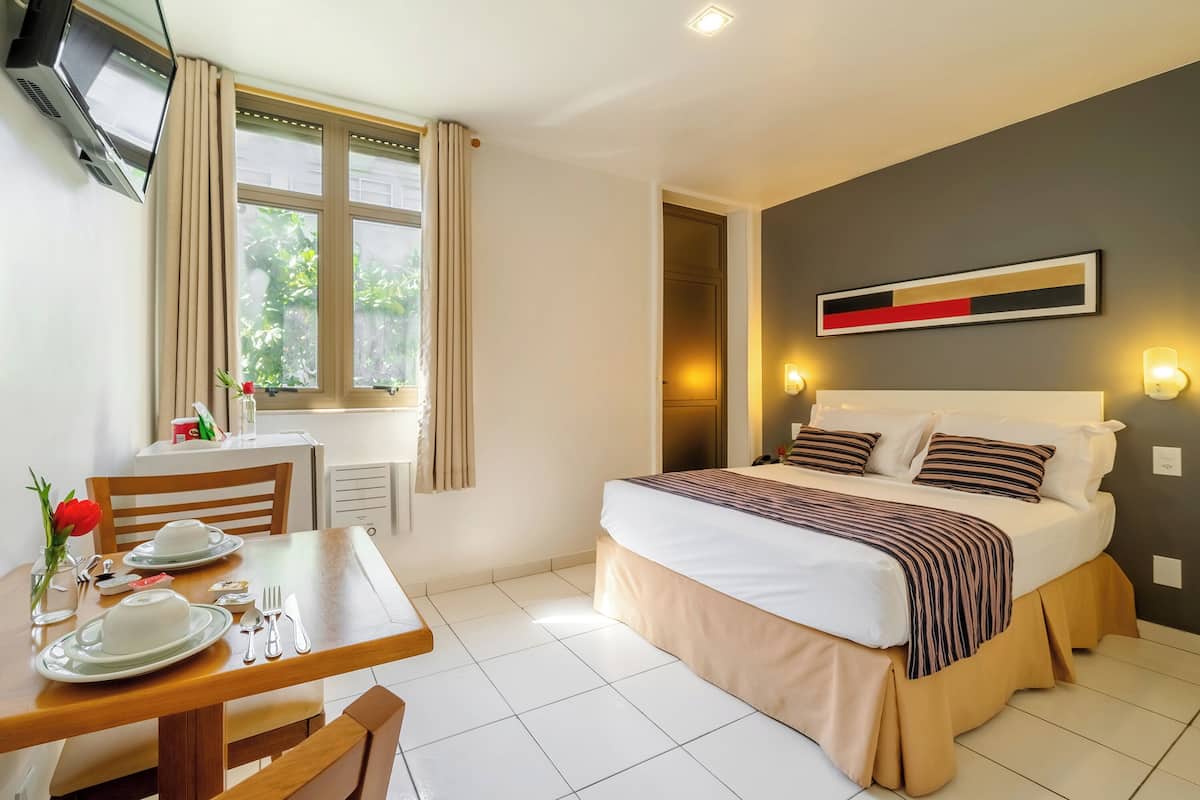 Ein Hotelzimmer mit Bett, Esstisch mit Tellern und Besteck, einem an der Wand montierten Fernseher und einem Fenster mit Vorhängen.