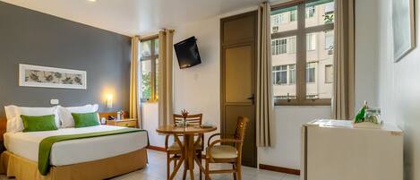 A hotel room with a bed, a dining table with chairs, a TV mounted on the wall, and a refrigerator.