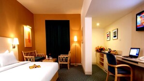 Quarto de hotel com cama, escrivaninha, cadeira e televisão na parede.