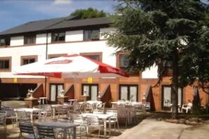 Un área de café al aire libre con mesas y sillas, un paraguas rojo y un edificio con varias ventanas.