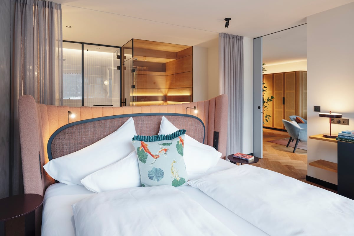 A modern hotel room with a neatly made bed, a small bedside table, a desk with a chair, and a large mirror.
