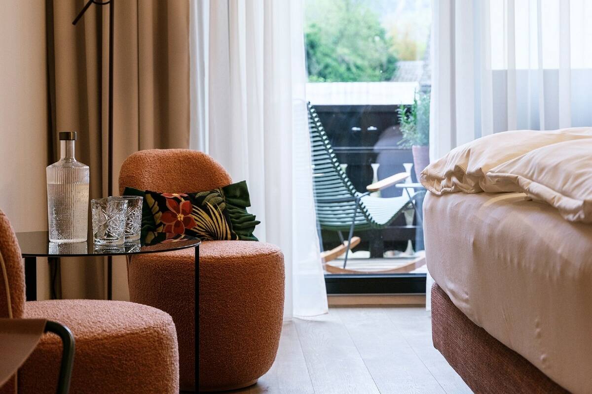 A bedroom with a bed, a chair, a table with a bottle and glasses, a floor lamp, and a view of greenery outside.