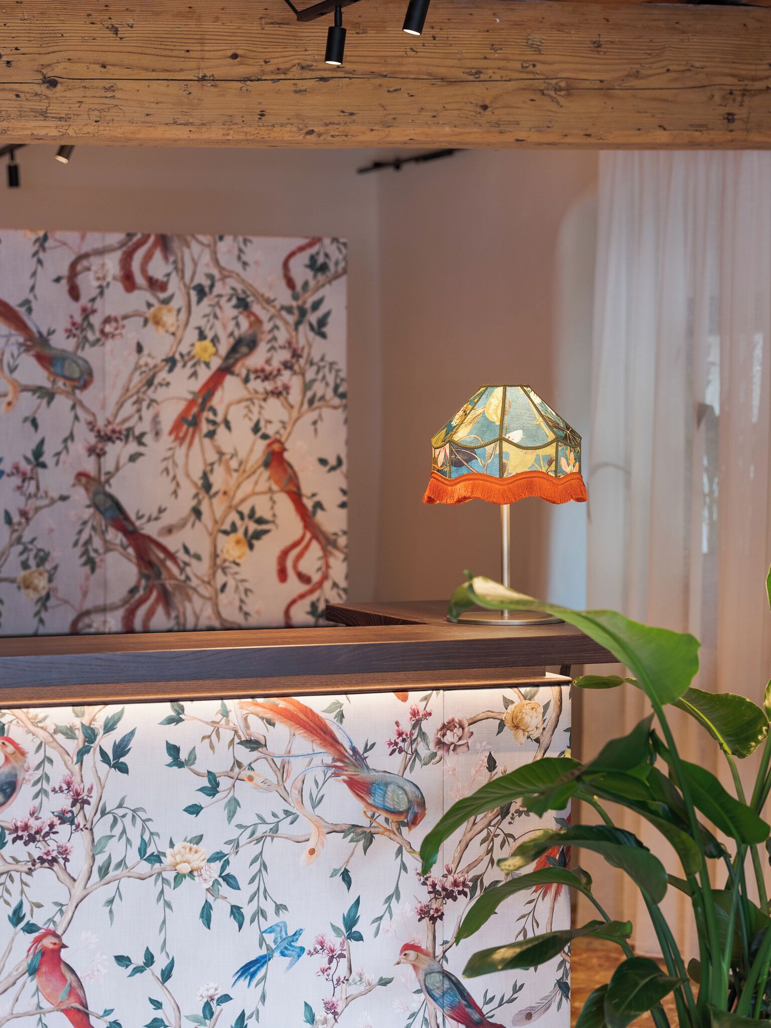 A room with a wooden ceiling, a wall with a bird-themed wallpaper, a table with a bird figurine, and a plant.