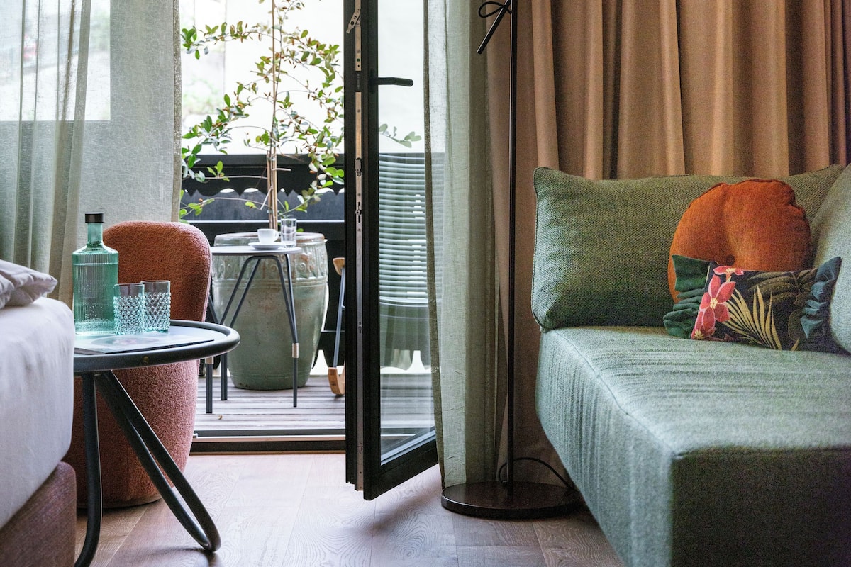 A hotel room with a bed, a green sofa, a floor lamp, a table with a glass, and a view of the outside.
