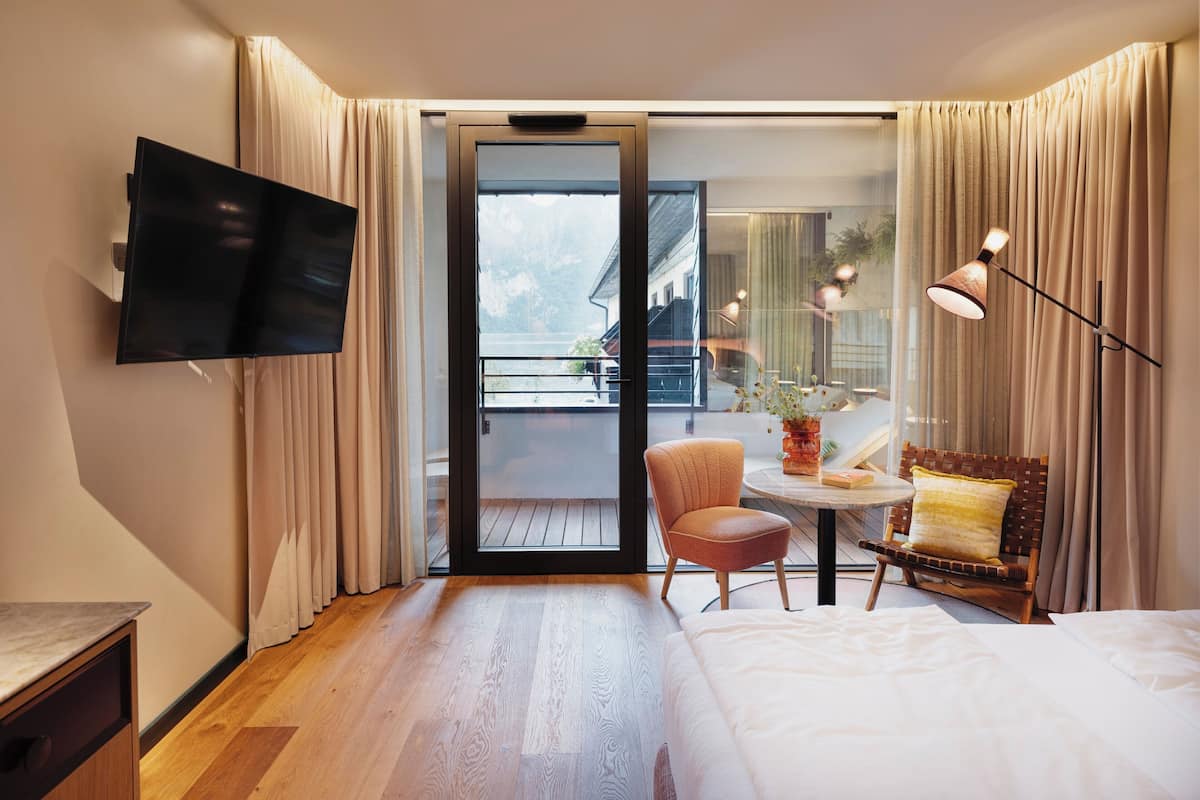 A hotel room with a bed, a TV mounted on the wall, a small table with a chair, and a balcony with curtains.
