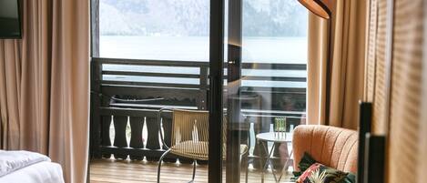 A hotel room with a wooden floor, a bed, a chair, a table, and a view of a balcony with a railing and a table.