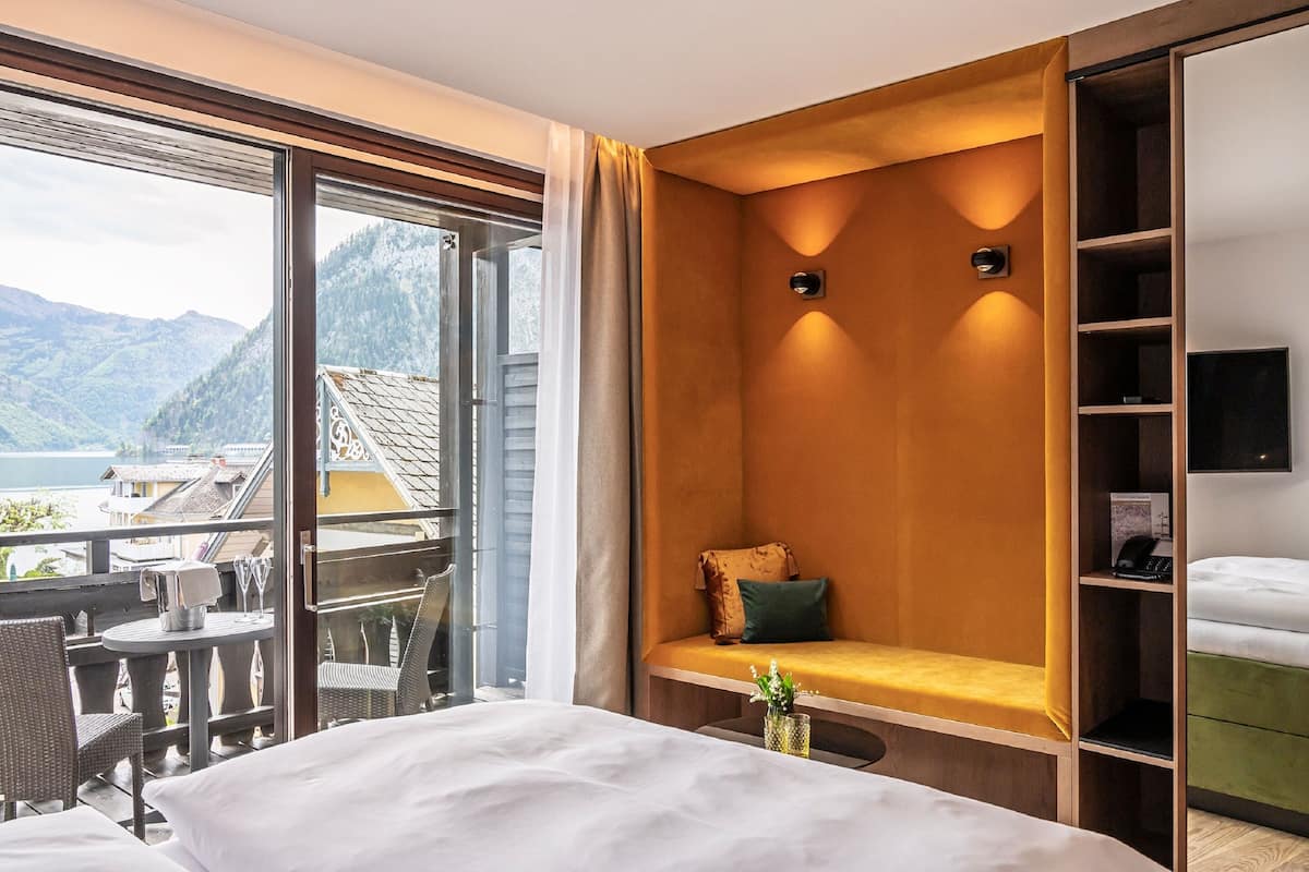 A hotel room with a large window, a balcony, a bed, a seating area, and a TV.