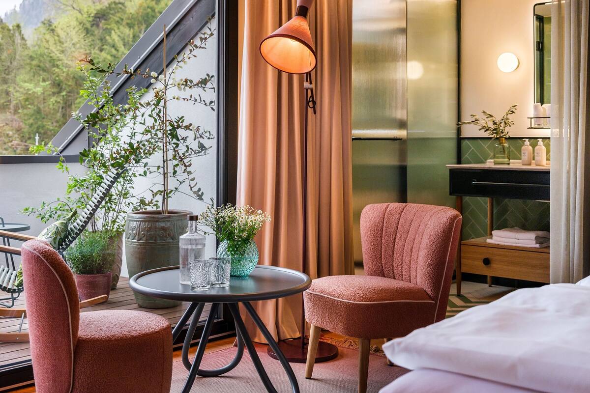 A modern hotel room with a balcony, a round table with chairs, a large window with curtains, a bedside table with toiletries, and a bed.