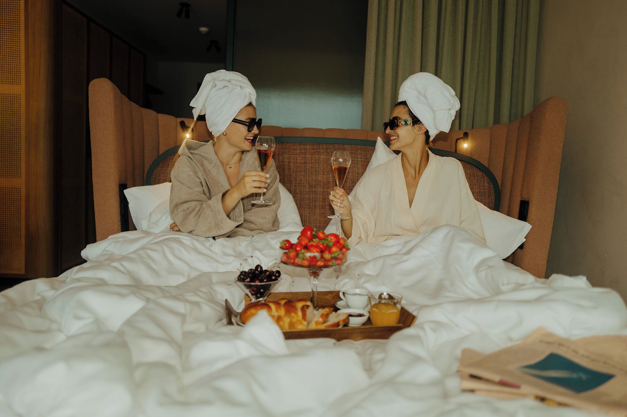 A hotel room with a bed, a tray with breakfast items, and two people sitting on the bed.
