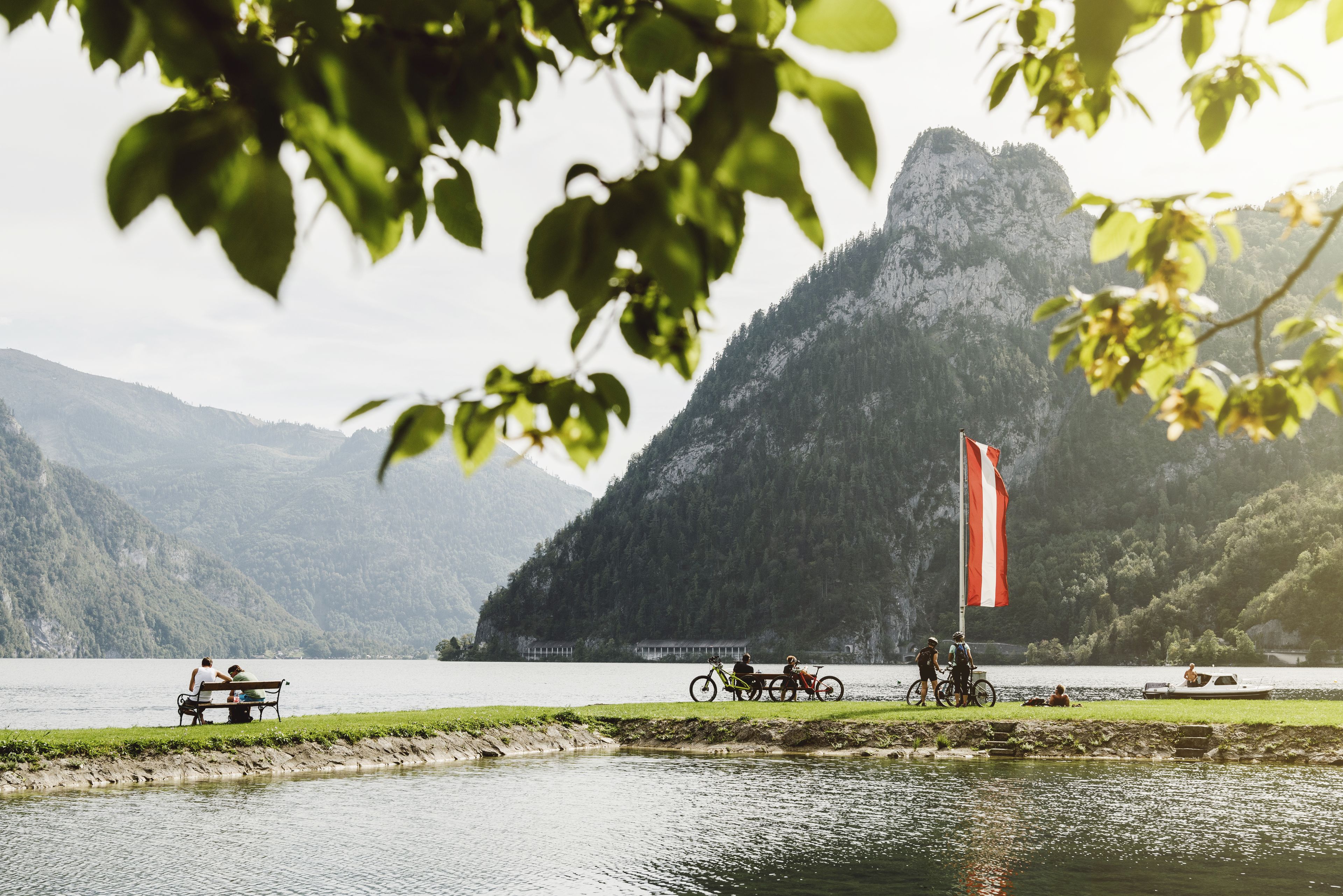 A lakeside view with a mountain in the background, a flag, and people on bicycles.