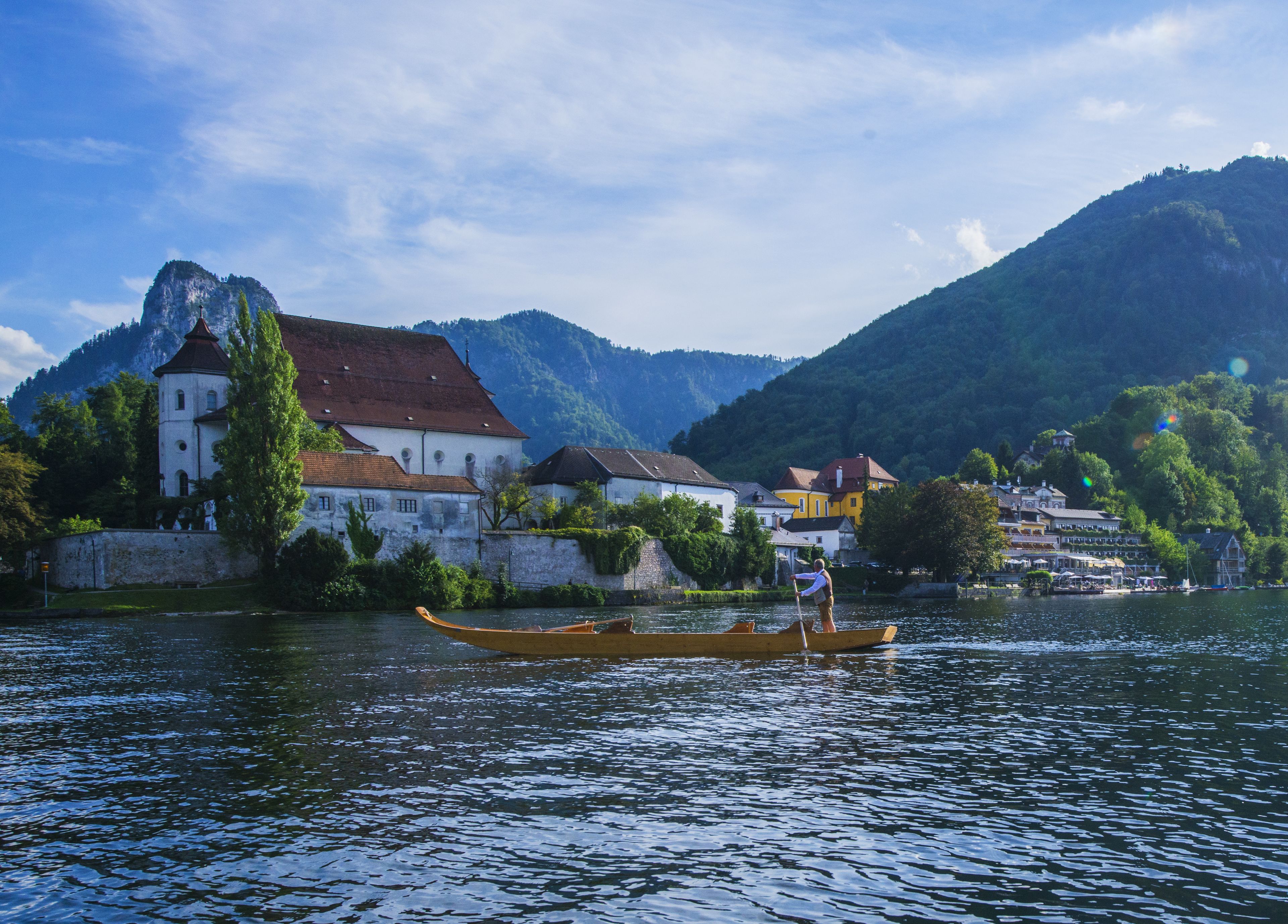 A historic building with a red roof, situated by a river with a person rowing a boat.