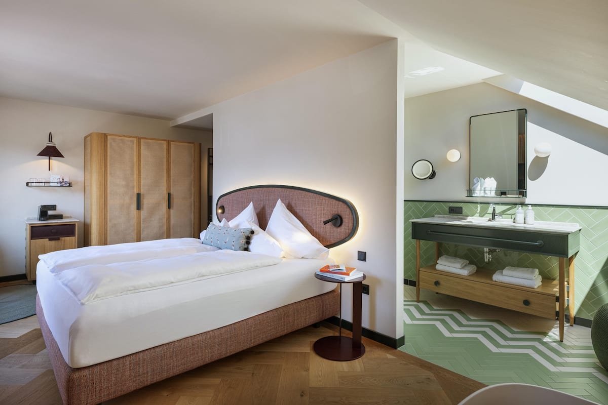A hotel room with a bed, bedside table, wardrobe, and a bathroom with a sink, mirror, and towel rack.