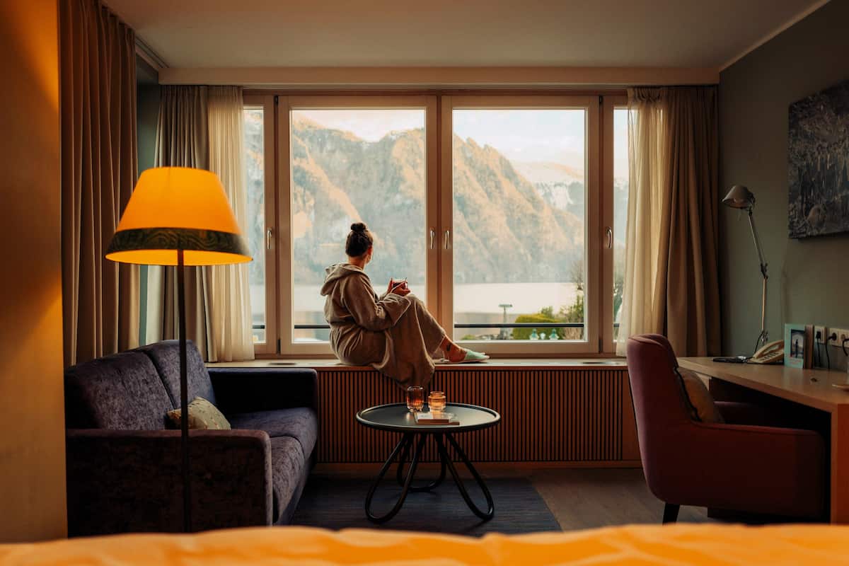 A hotel room with a large window, a sofa, a small round table, and a lamp.