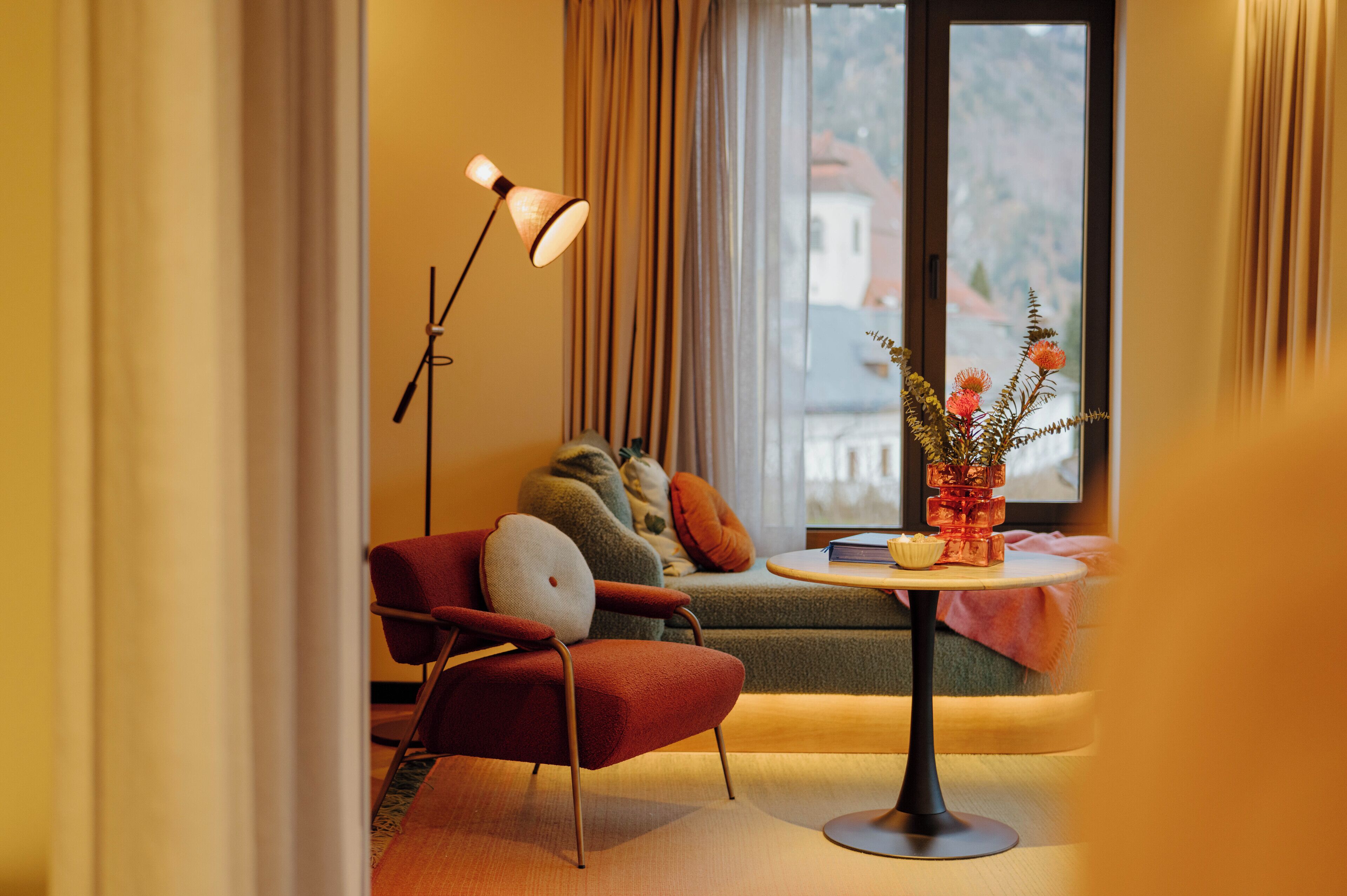Un salon chaleureux avec un fauteuil rouge, une petite table ronde ornée d’un vase de fleurs, un lampadaire et une vue sur une montagne par une grande fenêtre.