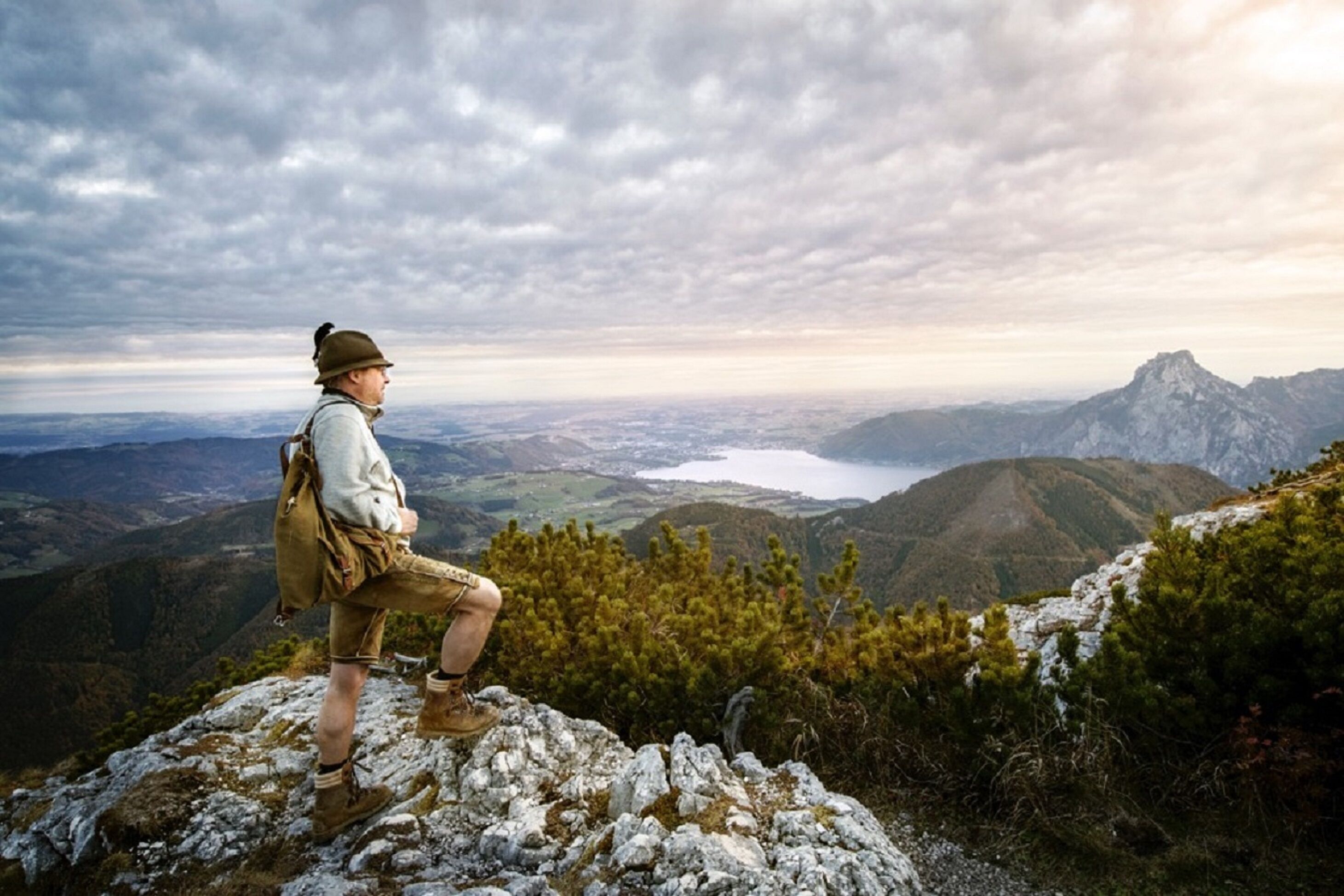 A person standing on a rocky outcrop overlooking a mountainous landscape with a lake and distant town.