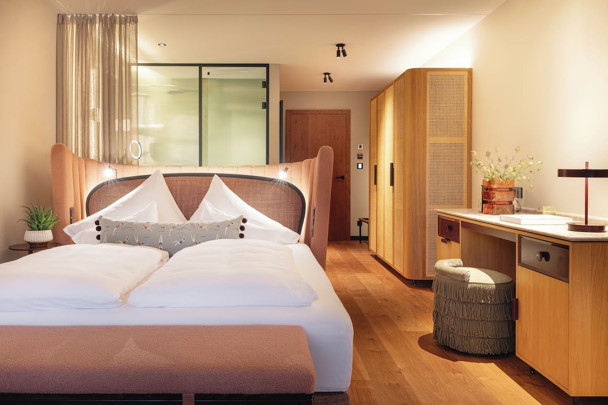 A modern hotel room with a large bed, a wooden wardrobe, a small stool, and a bedside table with a lamp.