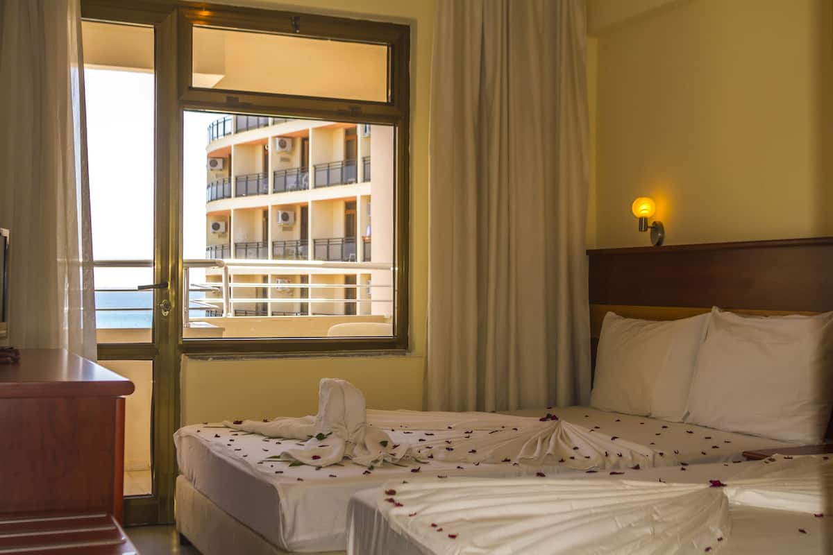 A hotel room with a bed, a wooden dresser, a window with curtains, and a view of a building with balconies.