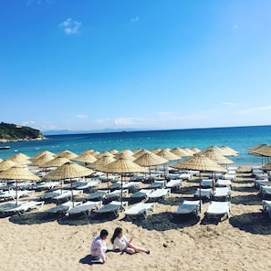 A beach with rows of sun loungers and thatched umbrellas, a clear blue sky, and a calm sea. - Hotel First Class (Didim)